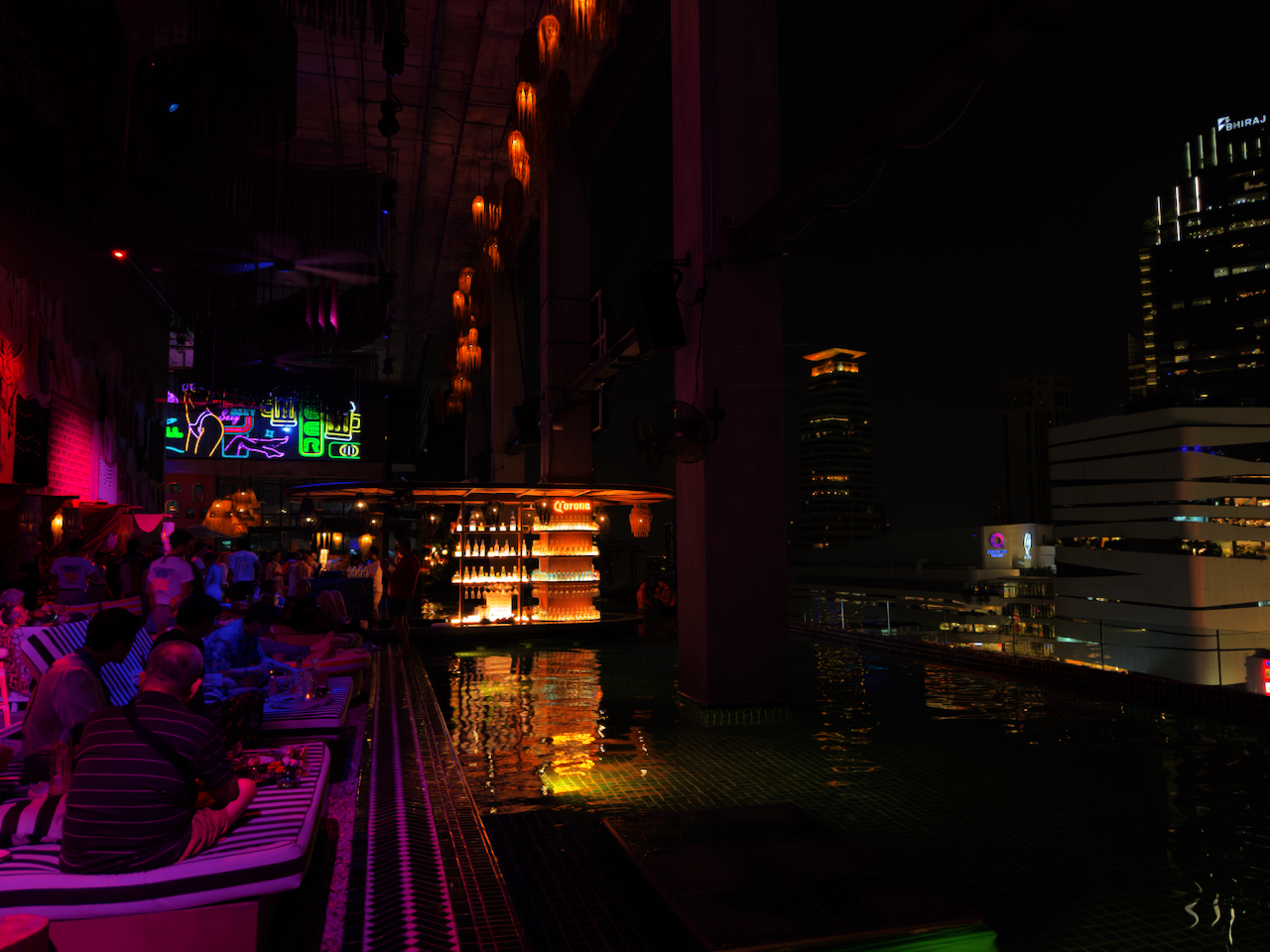 Rooftop-Pool-Bar in Thonglor bei Nacht – Gäste auf Liegestühlen, Infinity-Pool, Bangkoks Skyline im Hintergrund