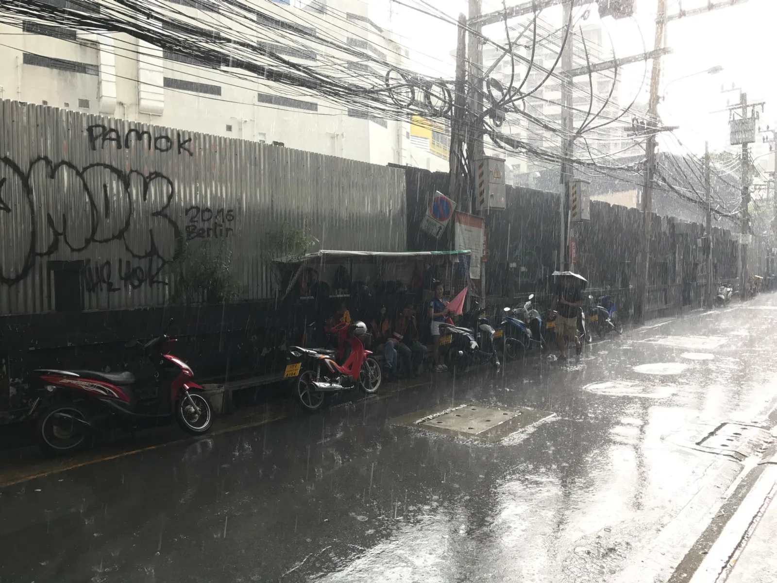 Tropischer Monsunregen prasselt auf eine Straße in Bangkok, Roller warten unter einem Vordach